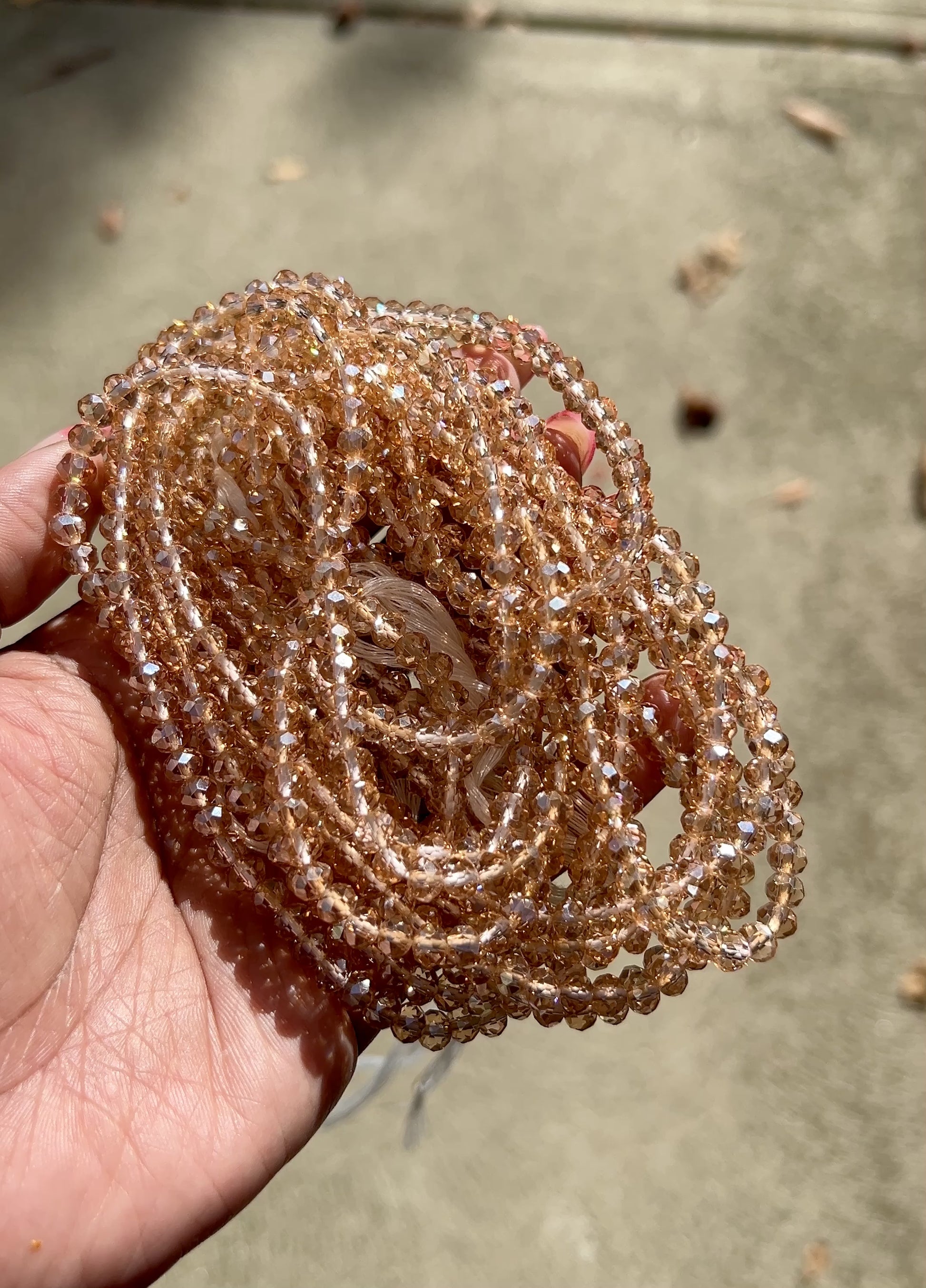 Rose gold waist beads made with faceted beads, coiled in a circular pattern in the palm of a hand on a cement background.