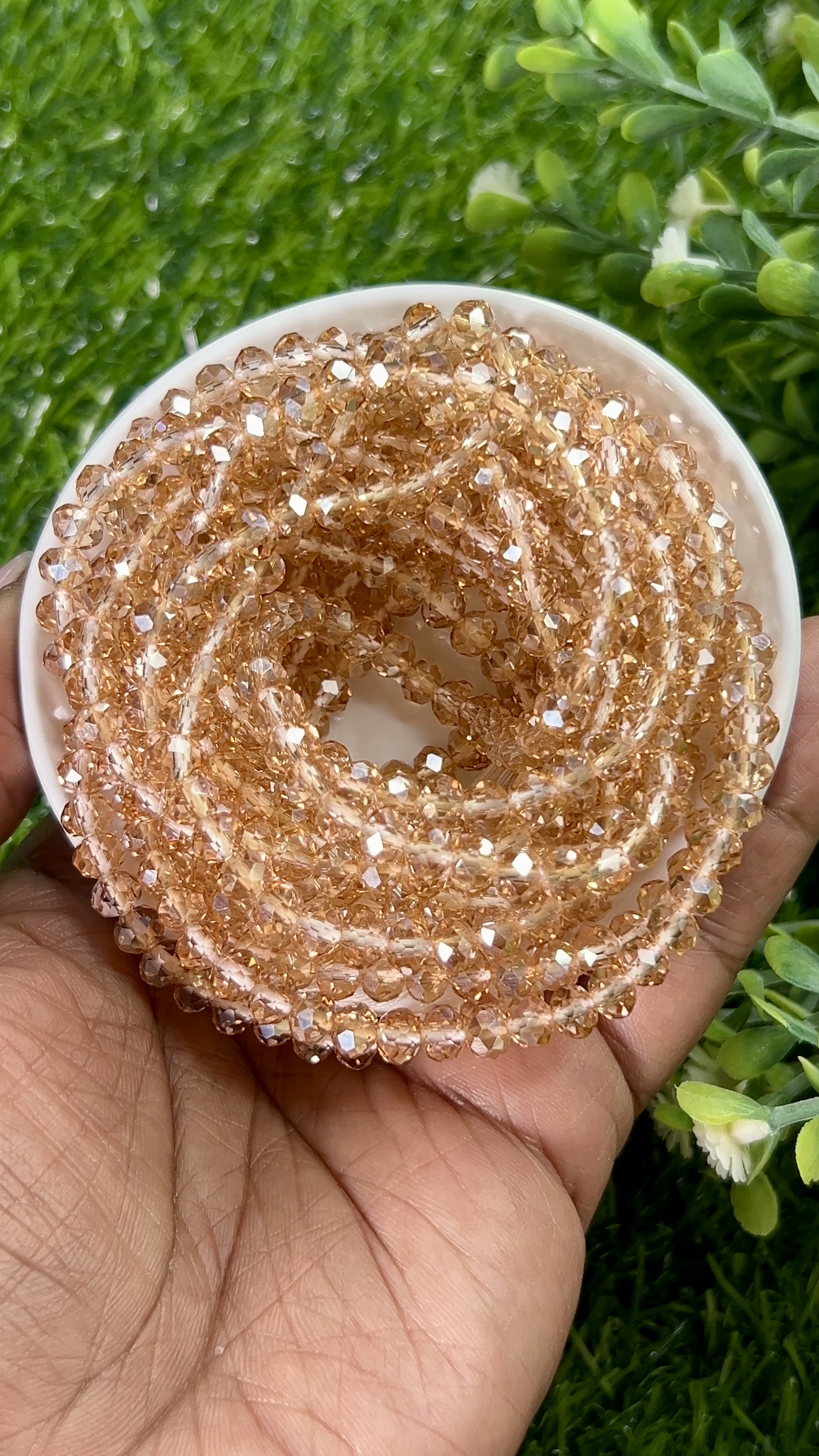 Rose gold waist beads made with faceted beads, coiled in a circular container on a grassy background.